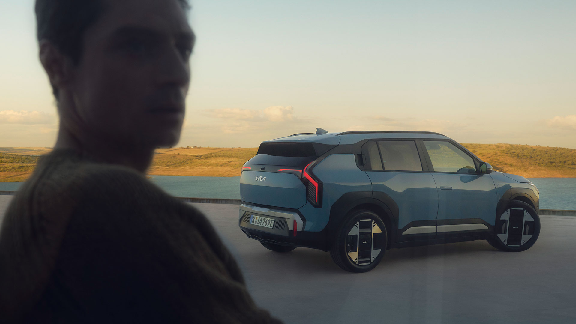 A man looks at a Kia EV3 parked by a serene body of water under a clear sky.