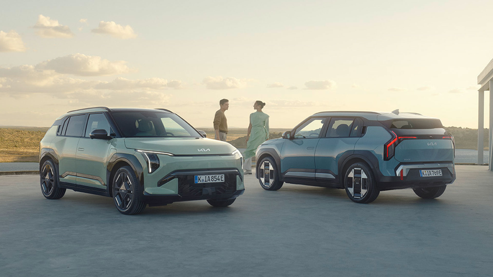 Two Kia EV3s parked in an open space with two people standing in the background against a scenic sky.